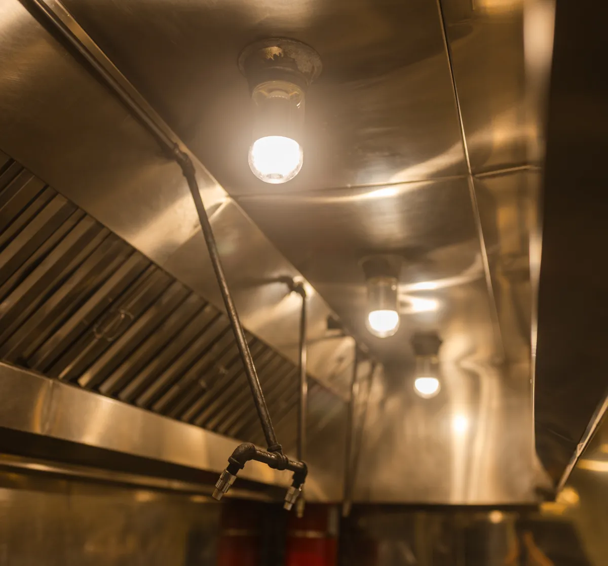 services-1 Close-up of industrial kitchen ceiling with metal vent hood, lights, and pipes.
