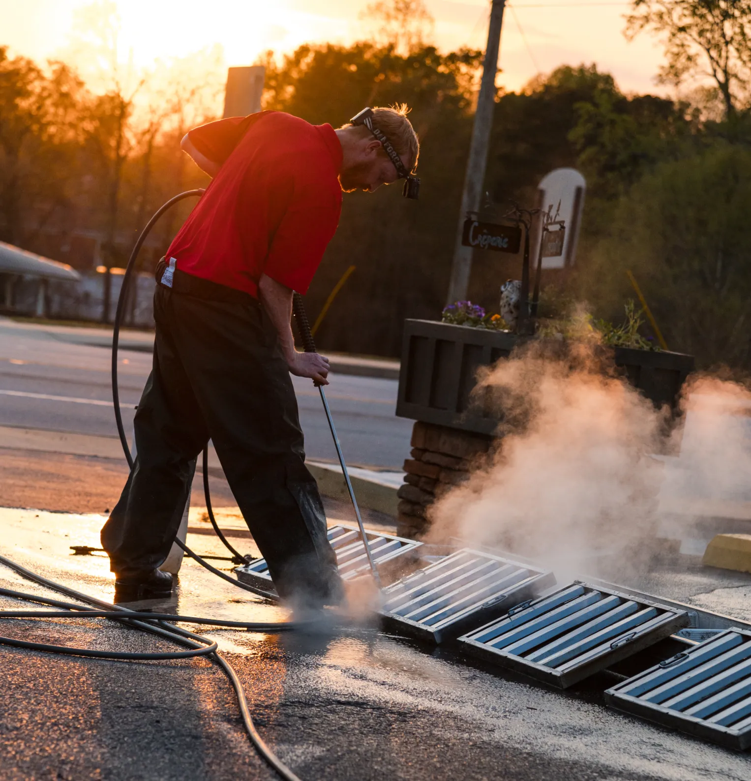 services-2 A man pressure washes metal grates on a sidewalk at sunset, with steam rising around him.